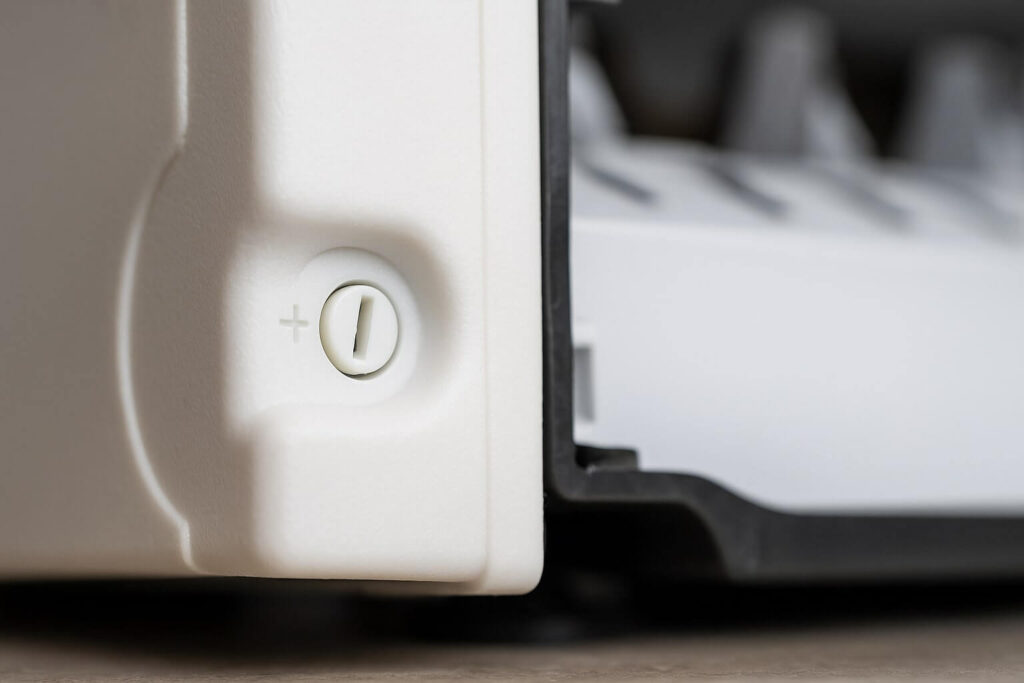 A close-up of a small plastic adjustment screw on the side of a modular refrigerator ice maker head, with '+' and '-' markings.