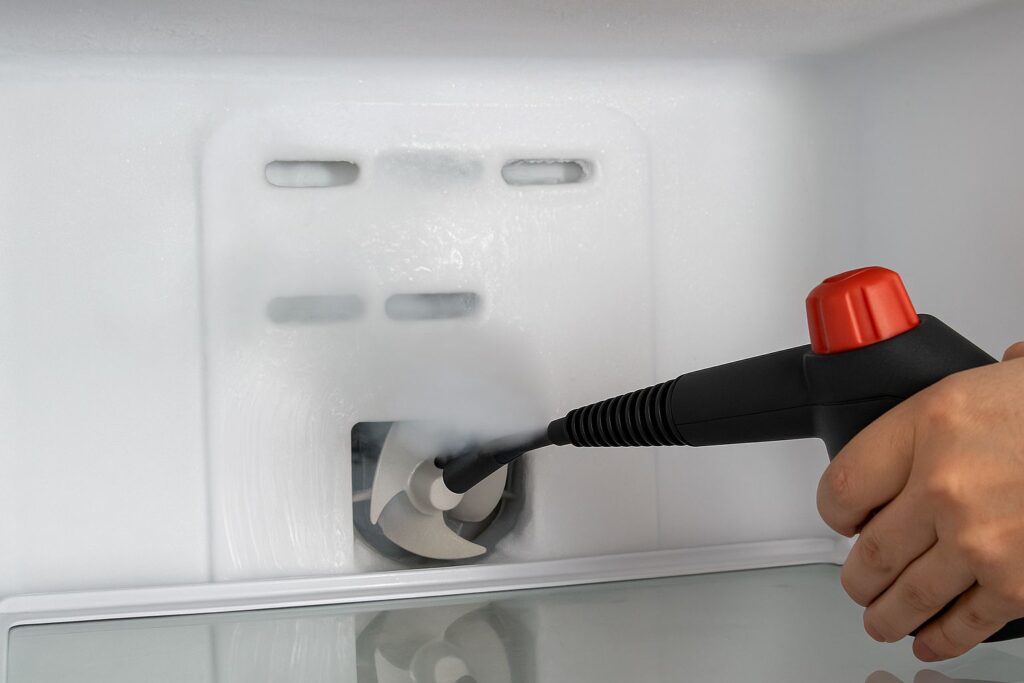 A photo showing a handheld steam cleaner nozzle pointing at the back wall of a freezer. Steam is gently billowing out, melting the ice around the air vents. This demonstrates the safe "manual defrost" method.