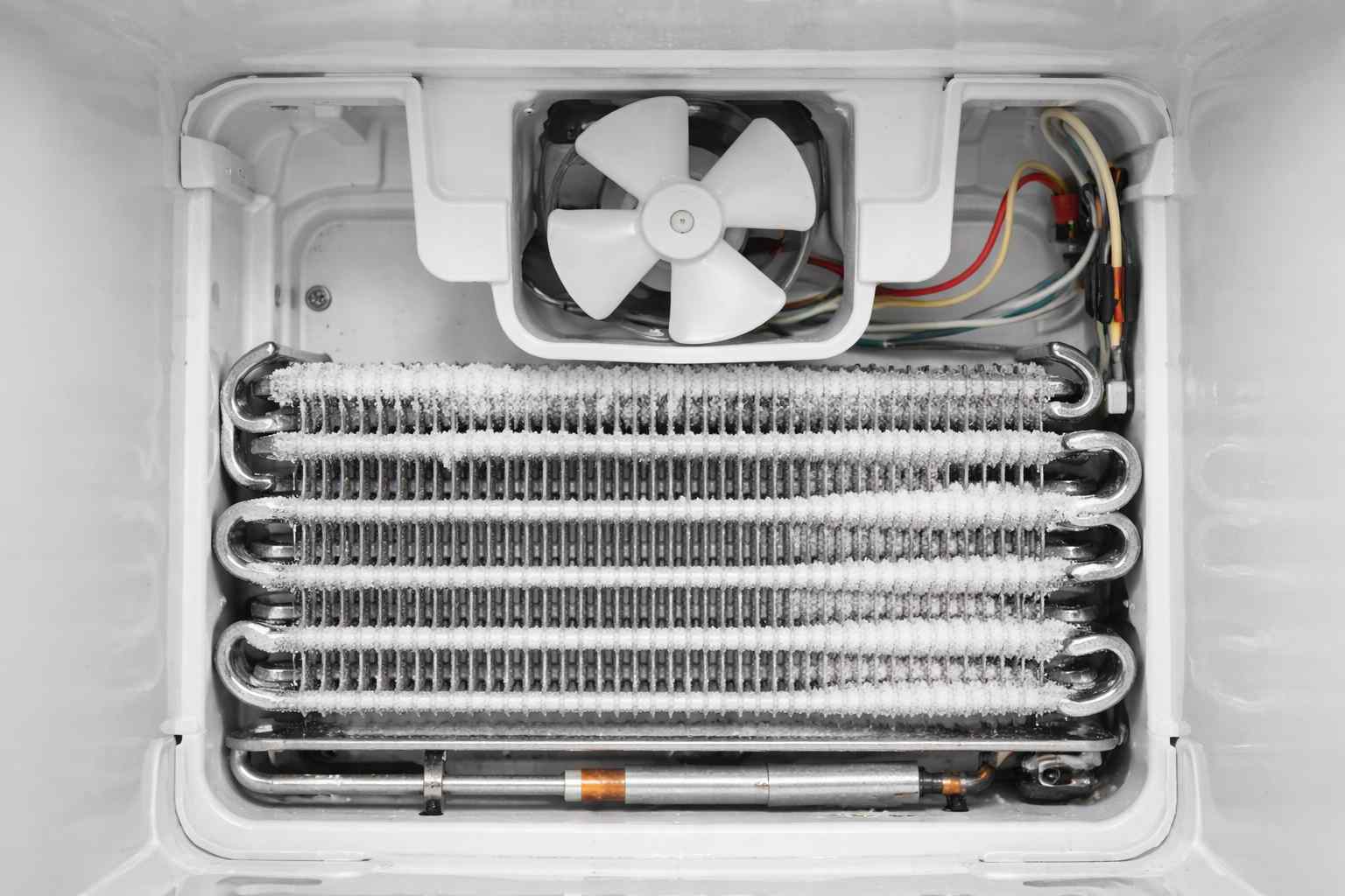 Looking directly into the back wall of a freezer compartment, exposing silver aluminum evaporator coils and a small plastic fan blade